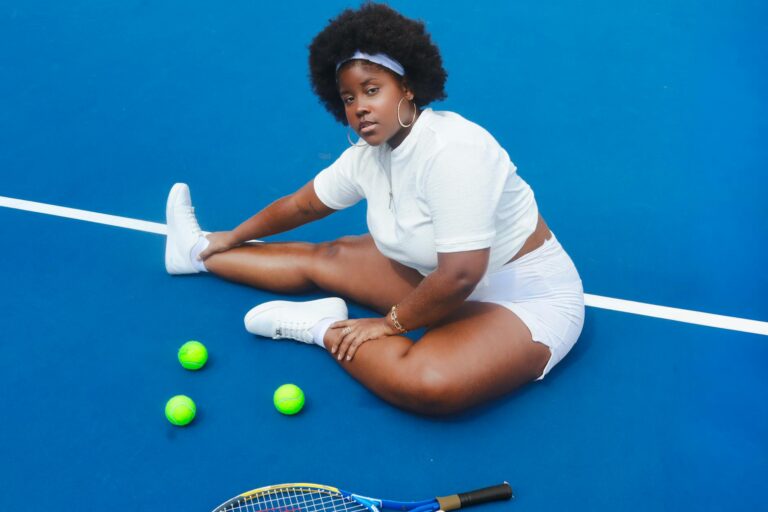 Confident young woman in athletic wear sitting on tennis court with racket and balls.