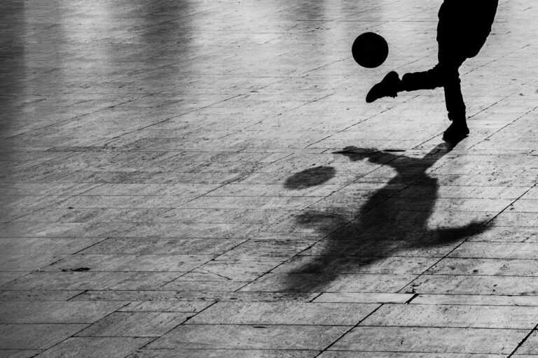 Shadow of a person kicking a football on a paved outdoor surface.