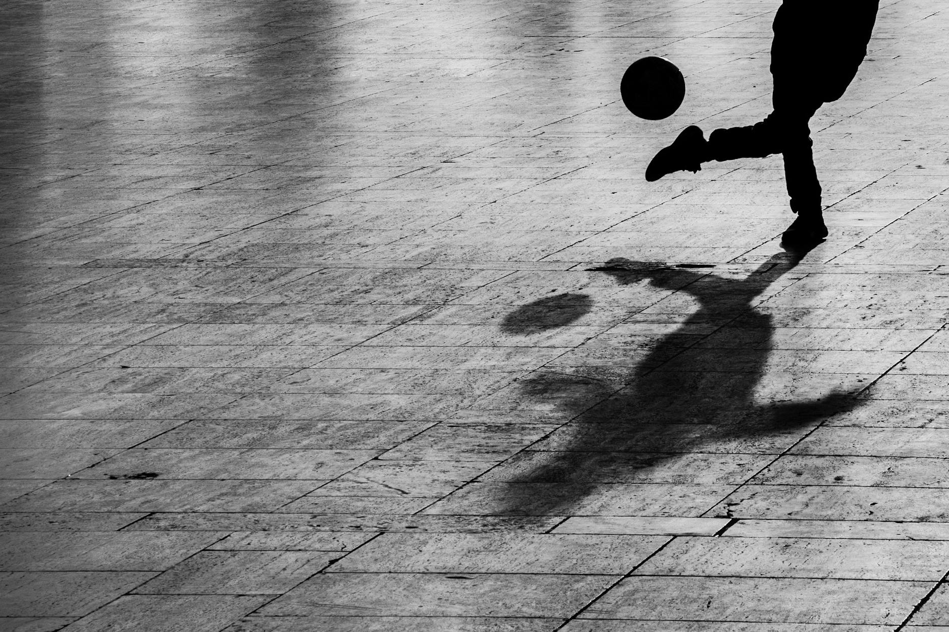 Shadow of a person kicking a football on a paved outdoor surface.