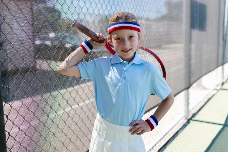 Smiling girl in tennis outfit standing by a fence with a racket on a sunny day.