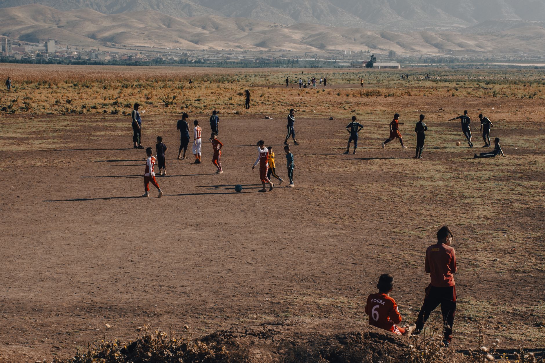 A group of children playing soccer in a rural field with mountains in the background. The scene captures a warm, outdoor ambiance.
