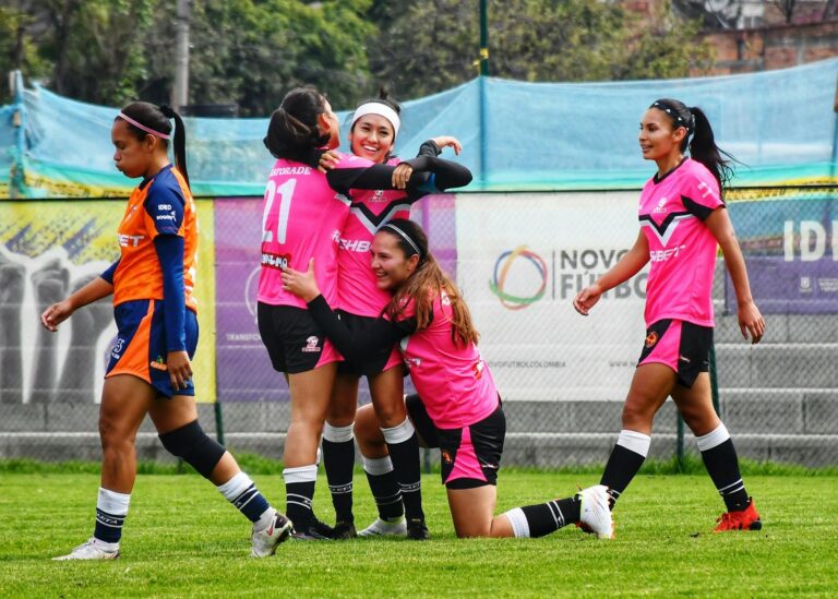A women's football team celebrating victory on the field with embraces and smiles.