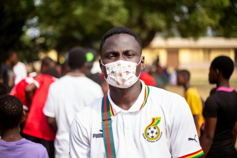 Young man wearing a mask at an outdoor event with a vibrant crowd in the background.