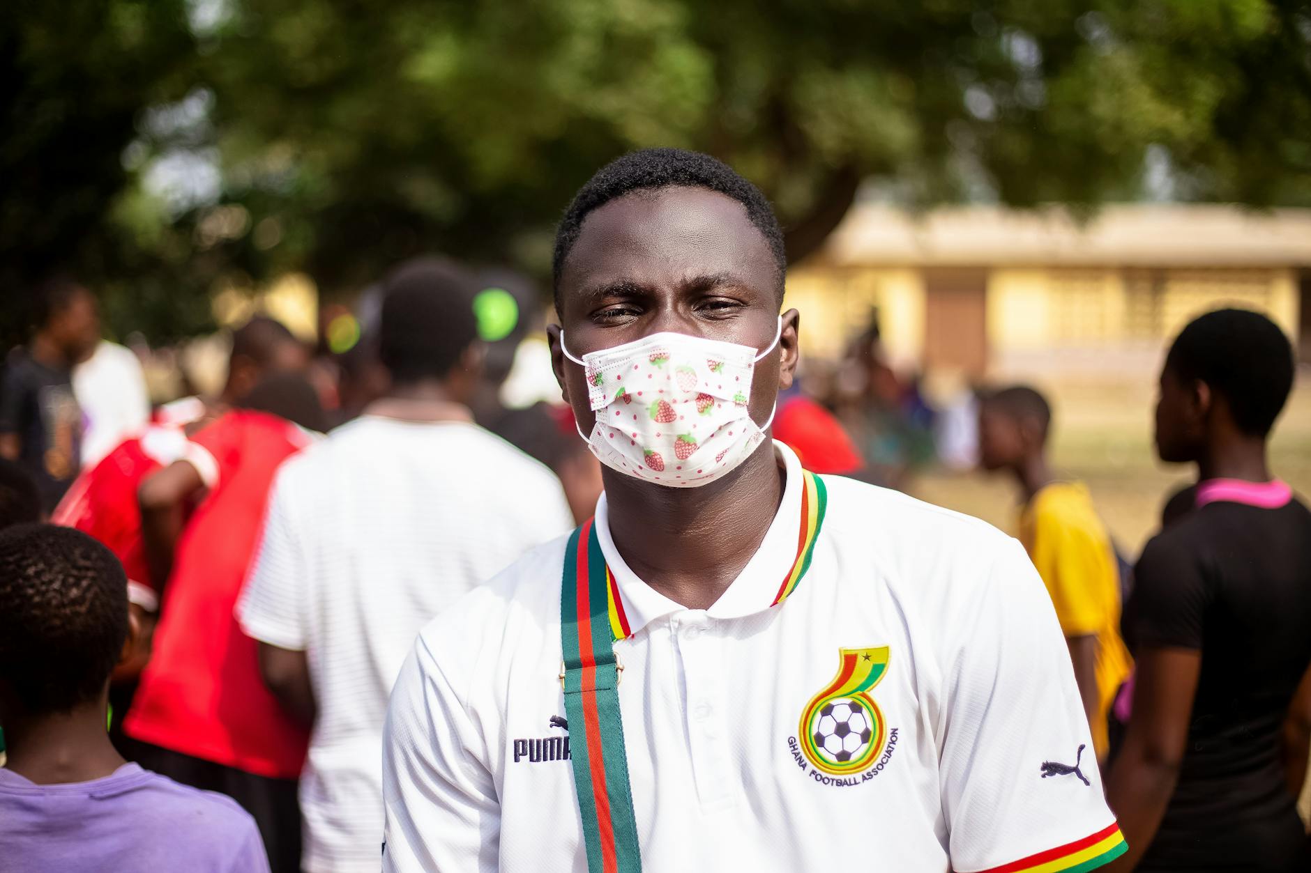 Young man wearing a mask at an outdoor event with a vibrant crowd in the background.