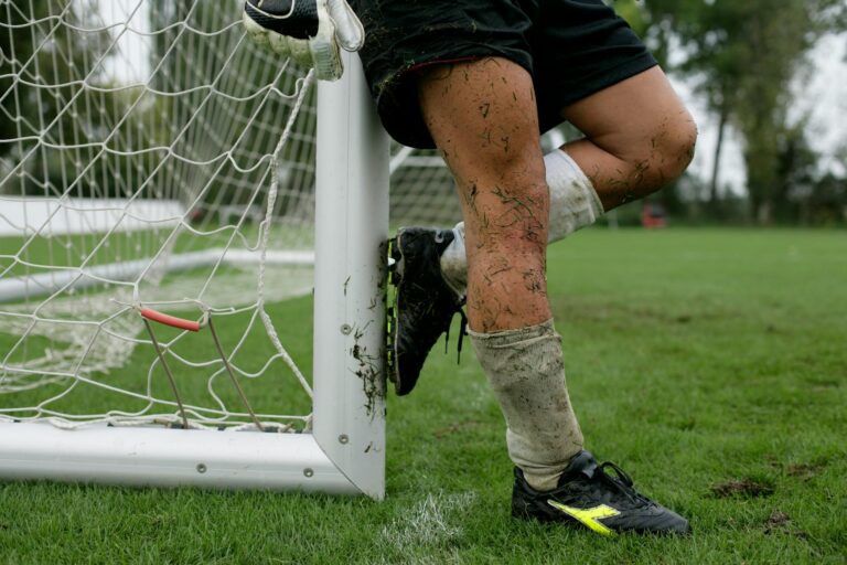 A soccer player with muddy legs leaning against a goal post on a grassy field.