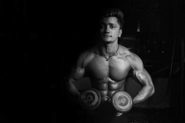 Shirtless male bodybuilder lifting dumbbells, showcasing muscular physique in a gym setting.