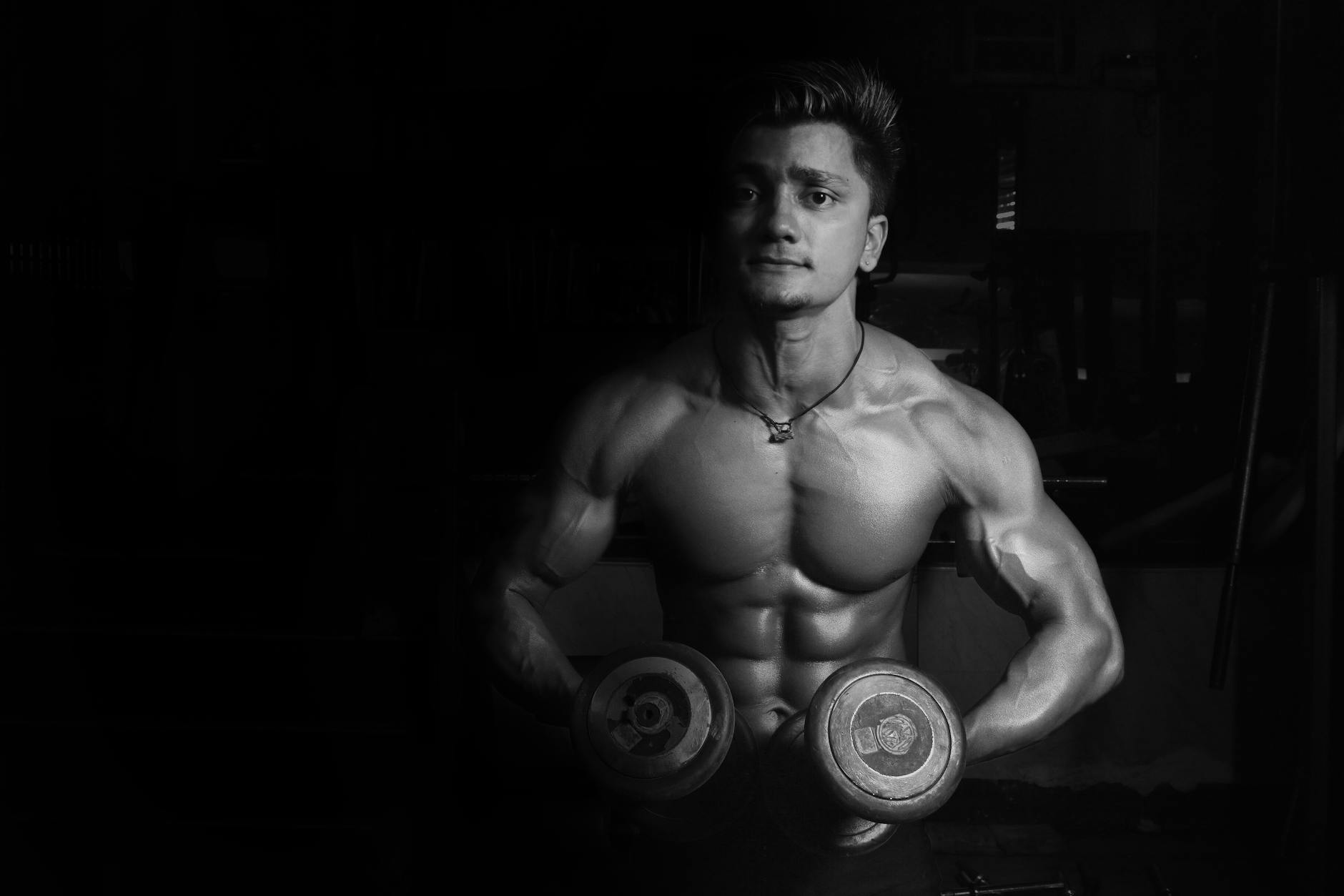 Shirtless male bodybuilder lifting dumbbells, showcasing muscular physique in a gym setting.