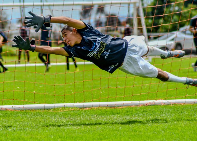 Soccer goalkeeper makes a dramatic save on a green field during a daytime match.