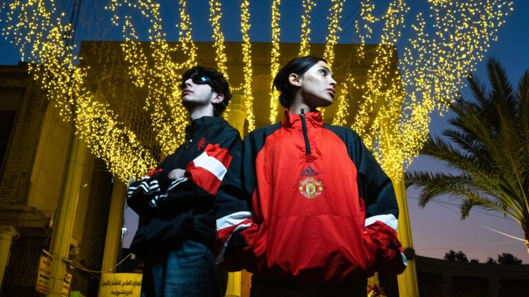 Two adults pose confidently under a canopy of glowing lights at night, wearing stylish jackets.