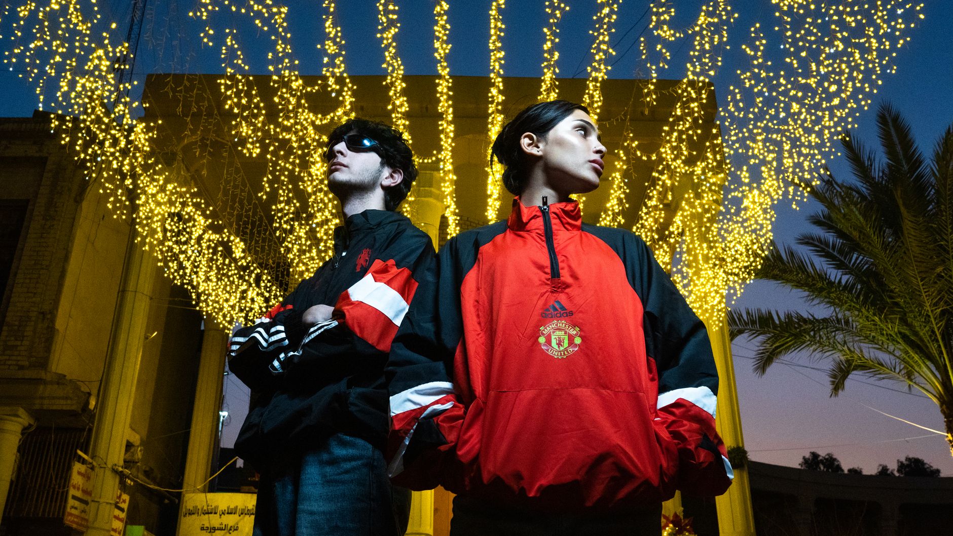 Two adults pose confidently under a canopy of glowing lights at night, wearing stylish jackets.