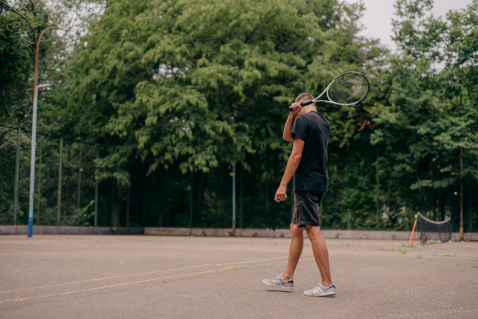 A young man holds a tennis racket while standing on an outdoor court surrounded by trees.