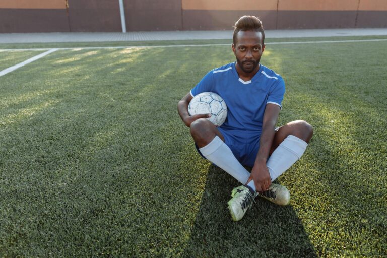 African man in blue jersey sitting on grass field holding a soccer ball.