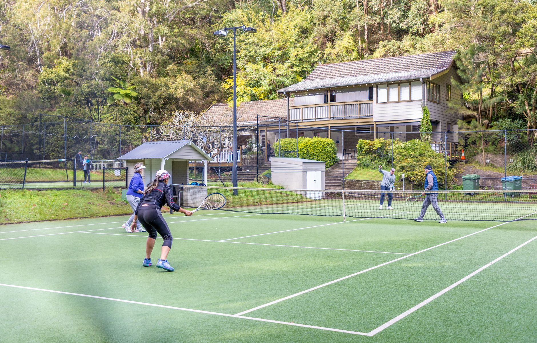 Four adults playing tennis on an outdoor court with a rustic house in the background.