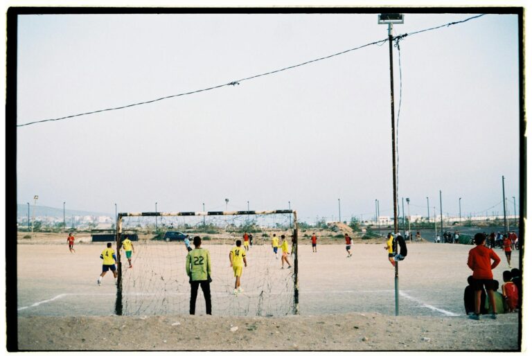 Players engaged in a soccer match on a dusty field in Morocco with scattered spectators.
