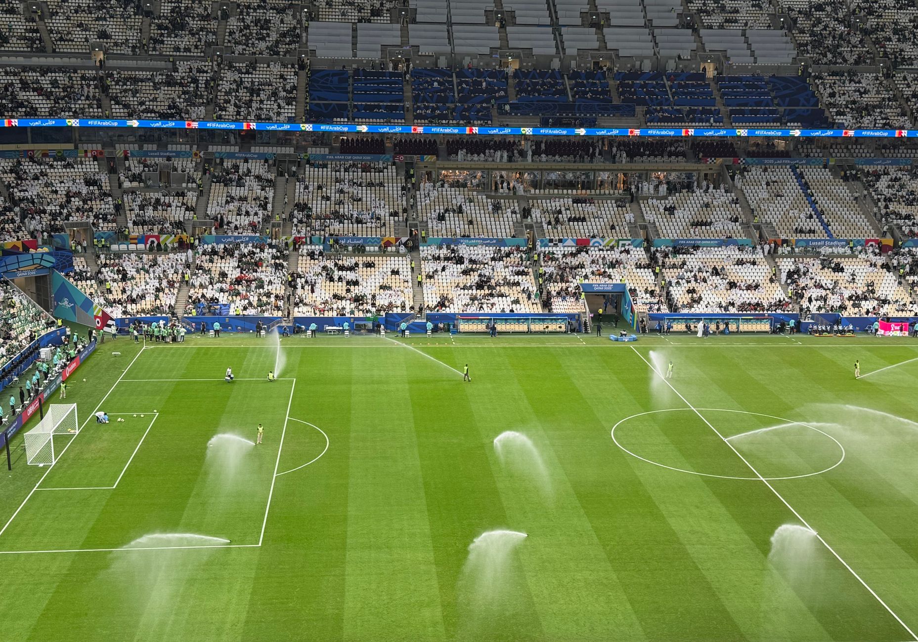 Soccer stadium field being watered with sprinklers under cloudy skies during daytime.
