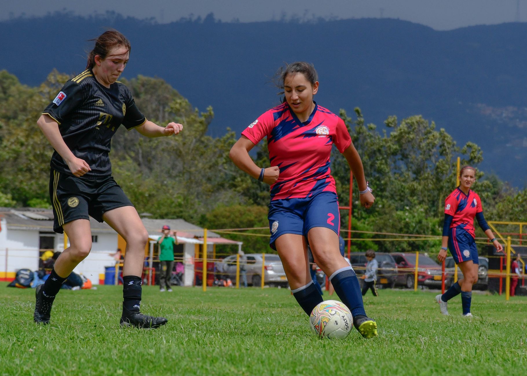 Women soccer players in action during a match on a lush green field with a mountain backdrop.