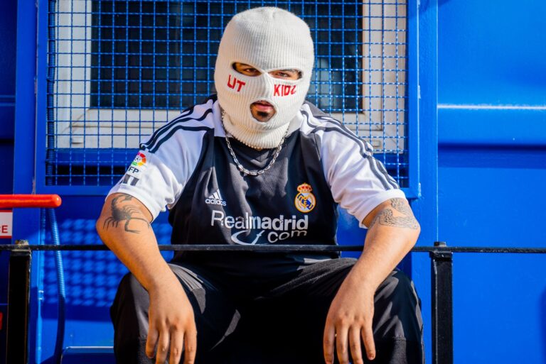 A person wearing a ski mask and sports jersey sits in an urban outdoor setting.