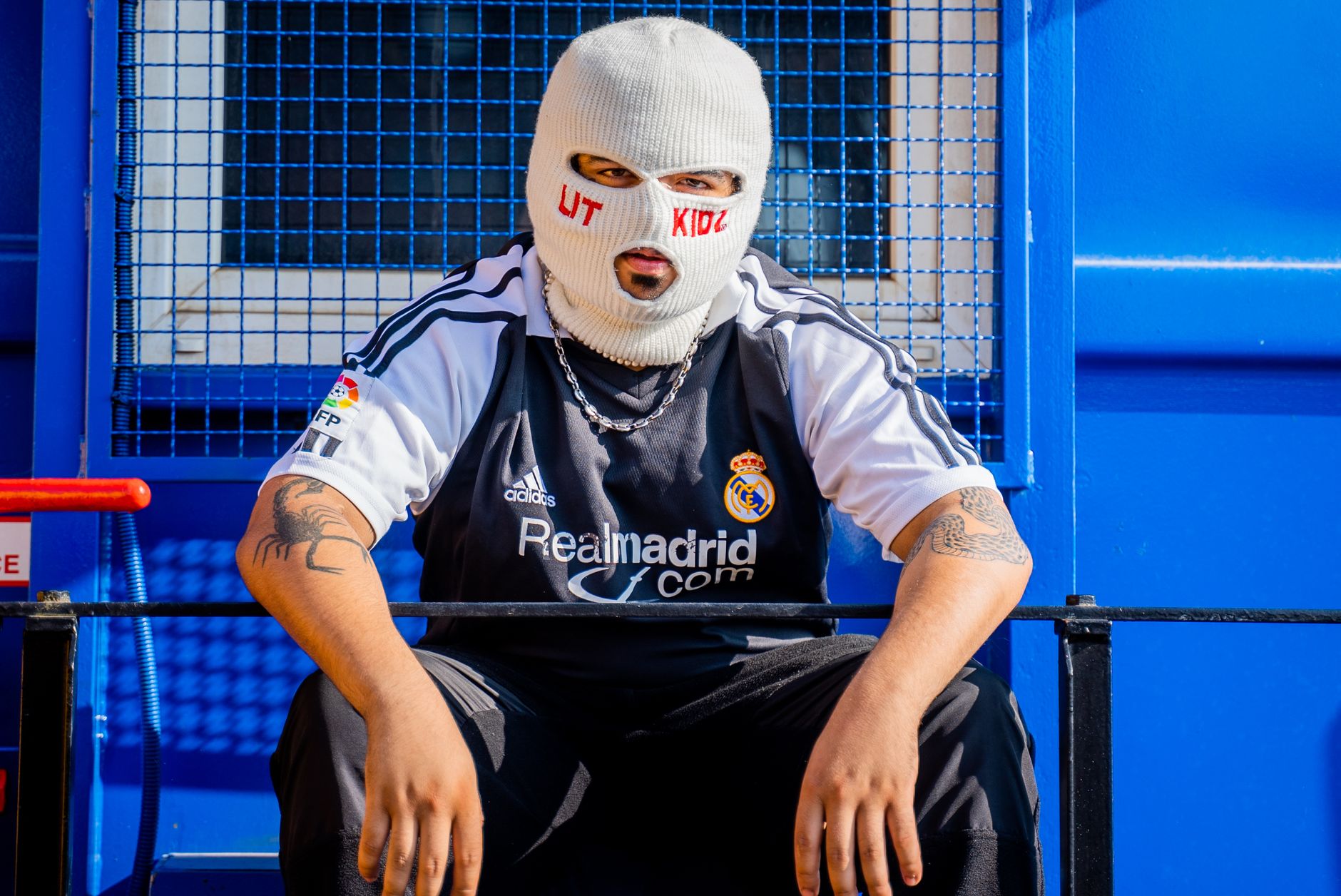 A person wearing a ski mask and sports jersey sits in an urban outdoor setting.