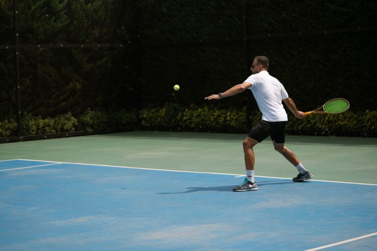 Tennis player hitting a backhand shot on a sunny outdoor court in Iran.