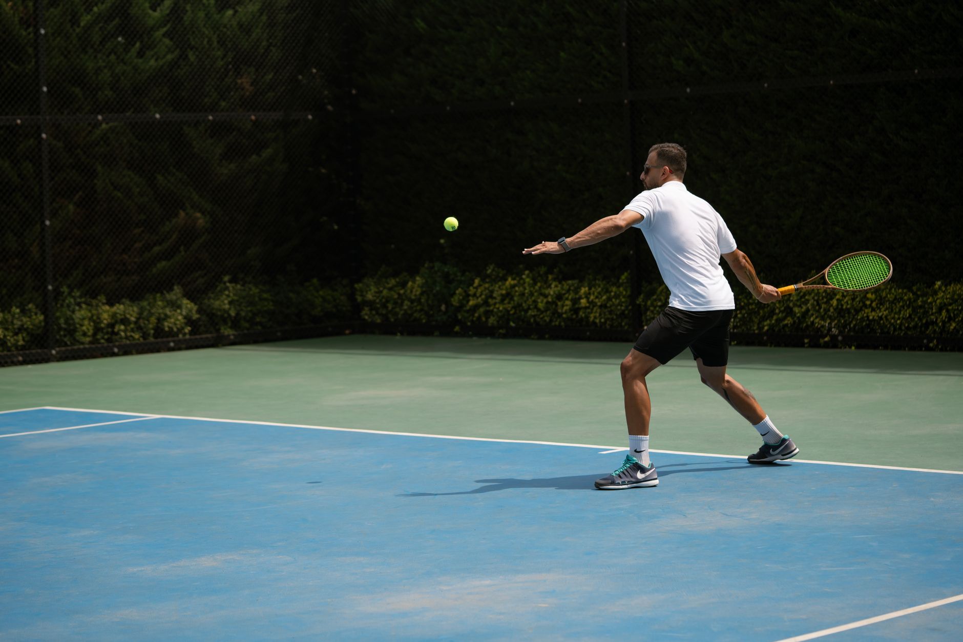 Tennis player hitting a backhand shot on a sunny outdoor court in Iran.