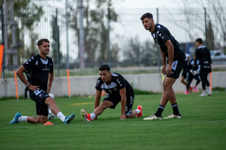 Three male football players stretching during training on a sunny field.