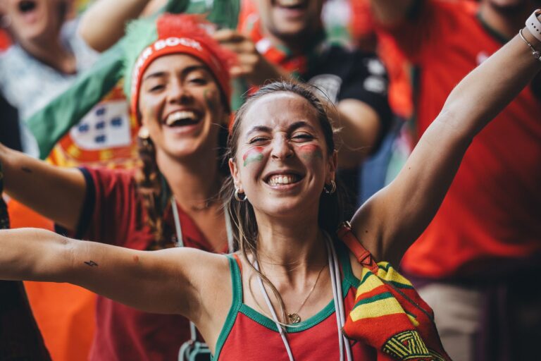 Excited women fans celebrate with flags and smiles during a sports event outdoors.
