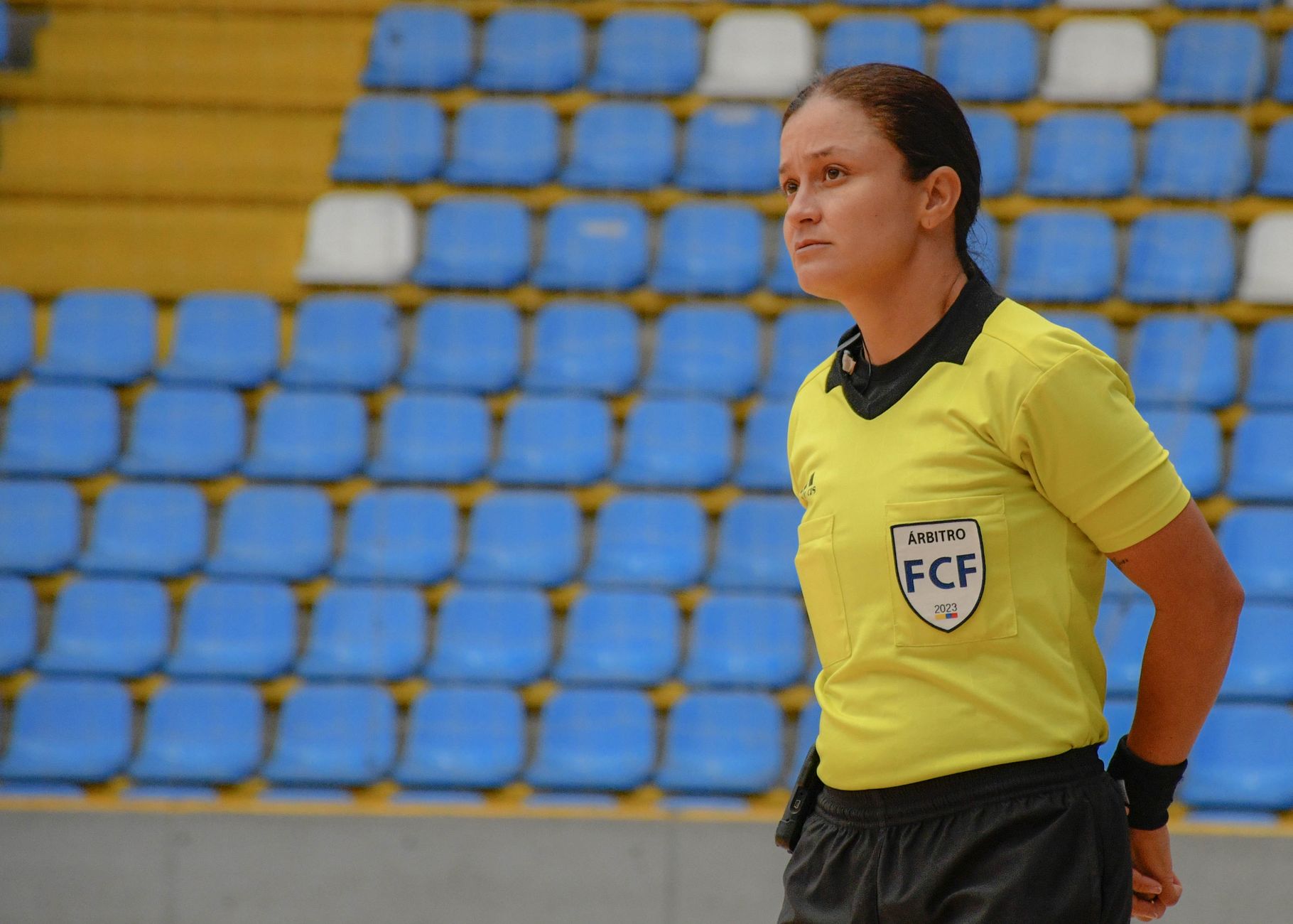 Woman futsal referee in yellow uniform stands focused in empty indoor stadium with blue seats.
