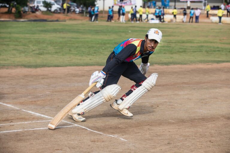 A cricket player in colorful attire bats during a daytime match on a sunny field.