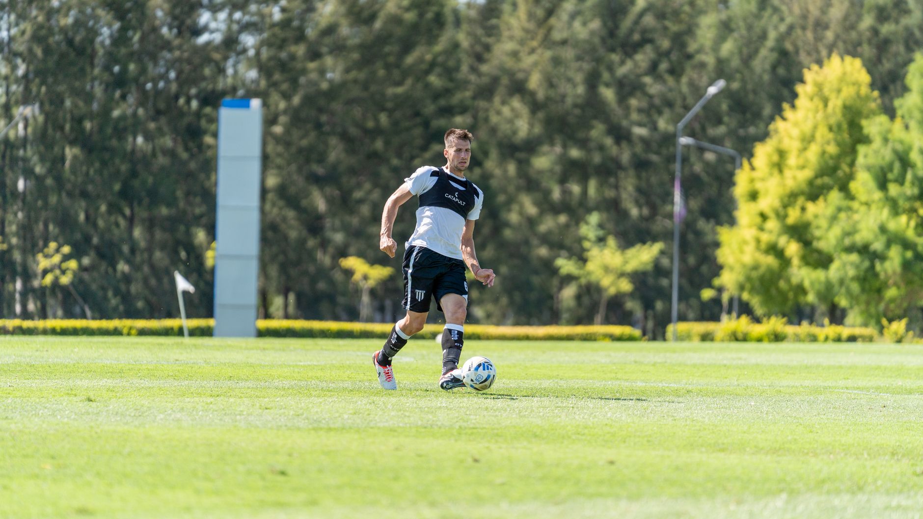 Soccer player dribbling a ball on a sunny outdoor field with lush greenery.