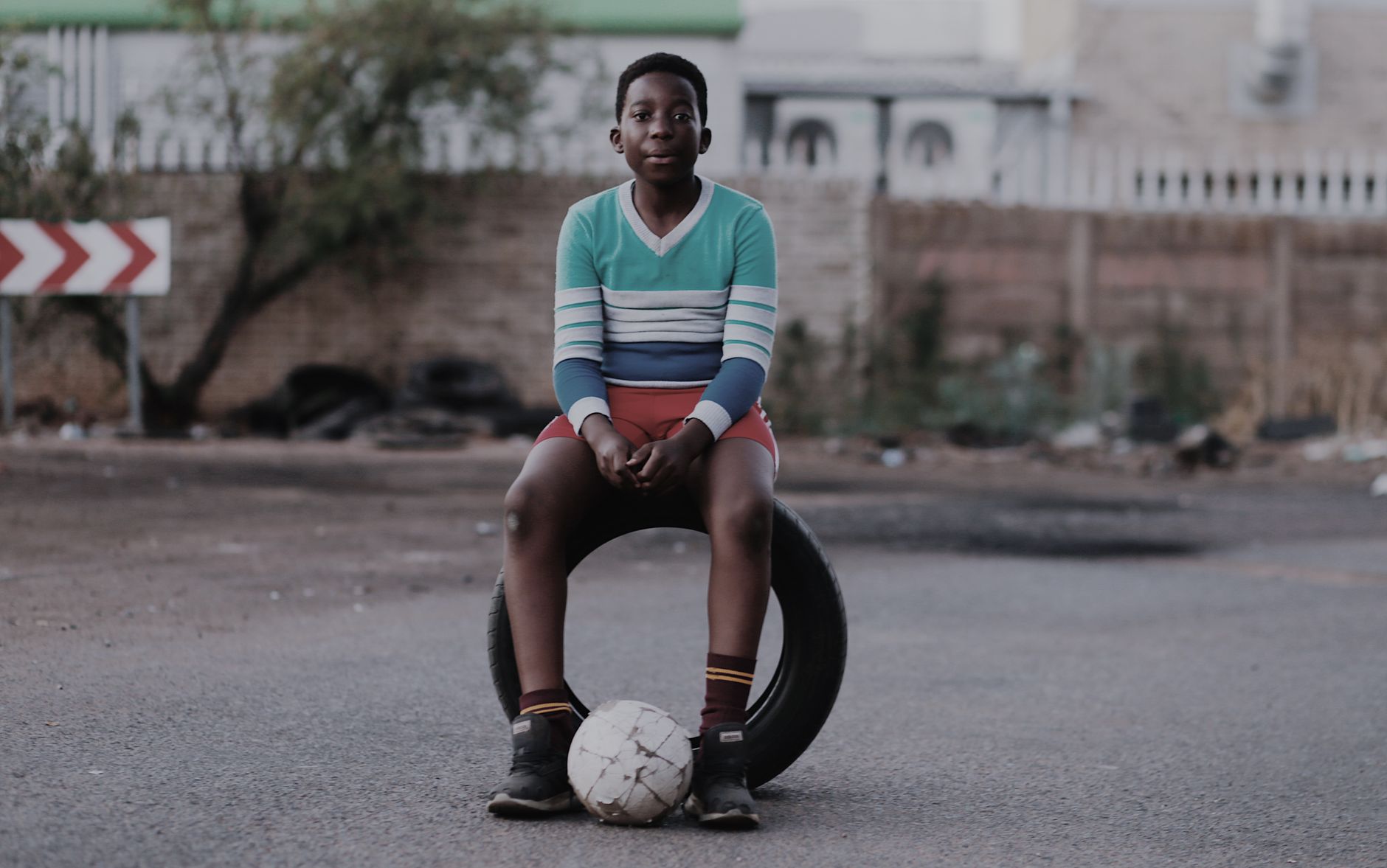 A young boy sits on a tire with a soccer ball in a quiet street, evoking a sense of play and solitude.