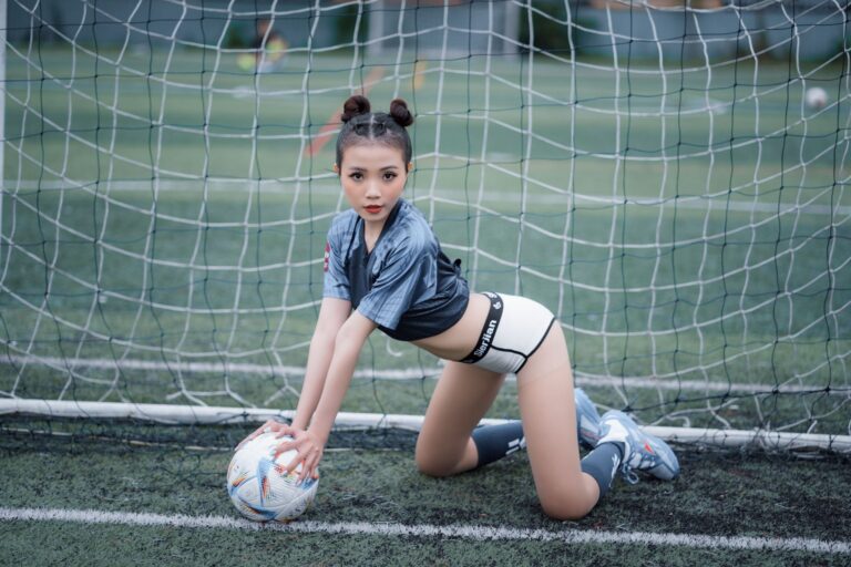 Fashion photoshoot of a woman in sportswear posing with a soccer ball on a football field.