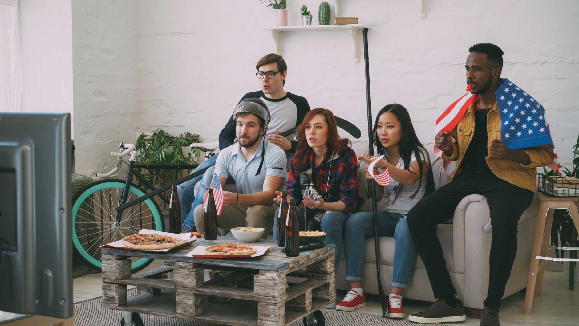 Diverse friends cheering for a sports game inside, surrounded by flags and snacks.
