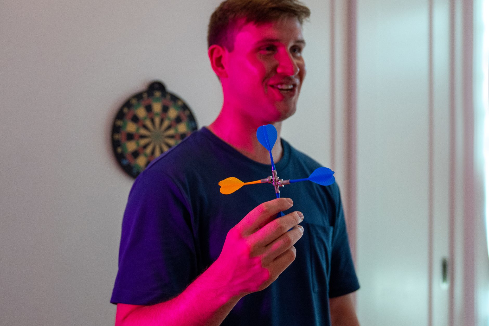 Man holding darts with vibrant pink lighting, smiling indoors near dartboard.