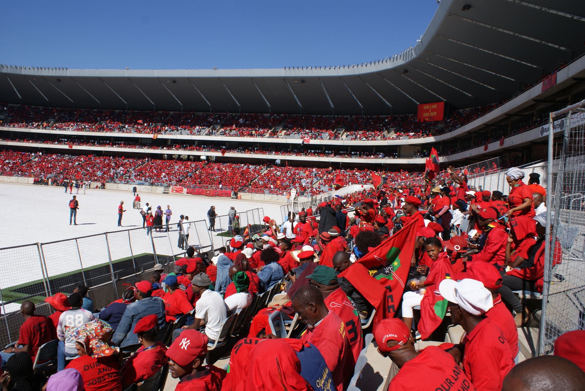 Vibrant crowd of sports fans in red cheering at a packed Soweto stadium under a clear blue sky.