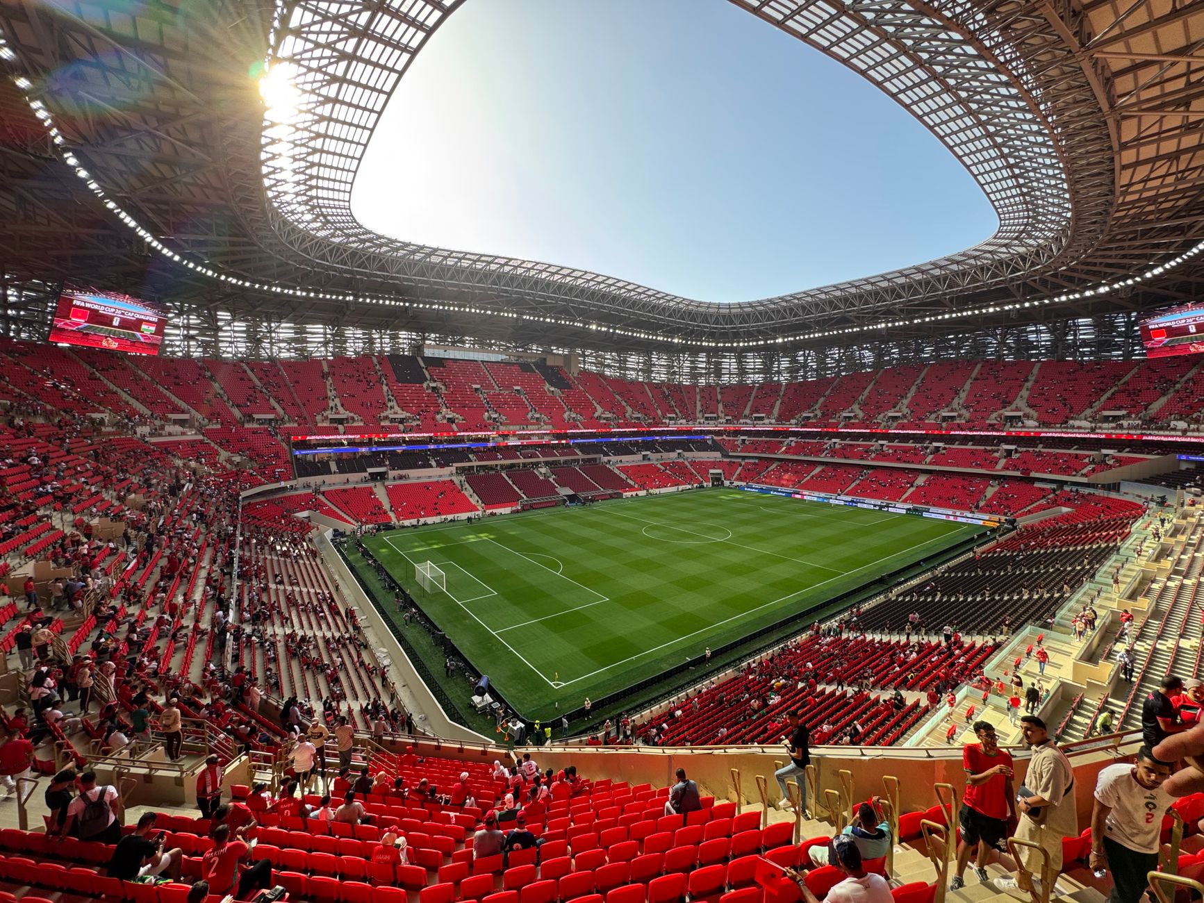 A modern football stadium with red seats, filled with fans during a daylight match, showcasing architectural design.
