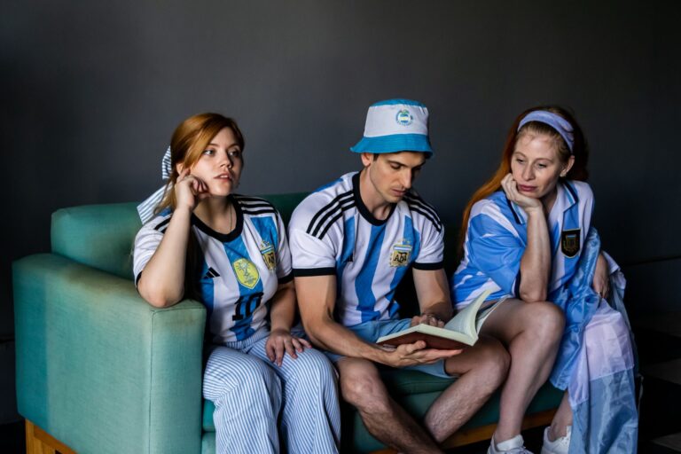 Three adults in Argentina soccer jerseys reading a book indoors on a couch.
