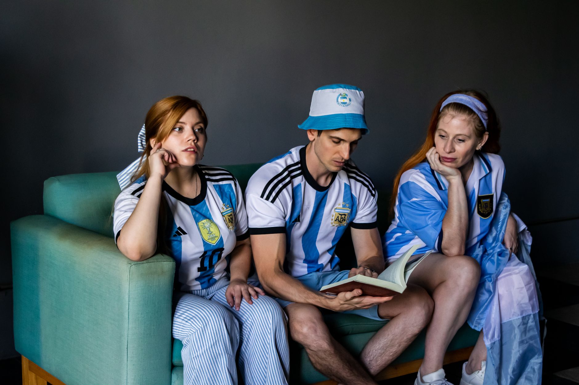 Three adults in Argentina soccer jerseys reading a book indoors on a couch.