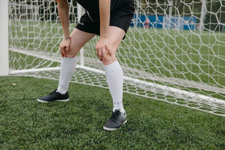 Soccer player resting in front of the goal on a grassy field, wearing sports uniform and sneakers.