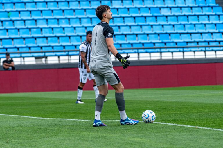 Goalkeeper holding a soccer ball during a match on a bright green field with empty blue seats in the background.