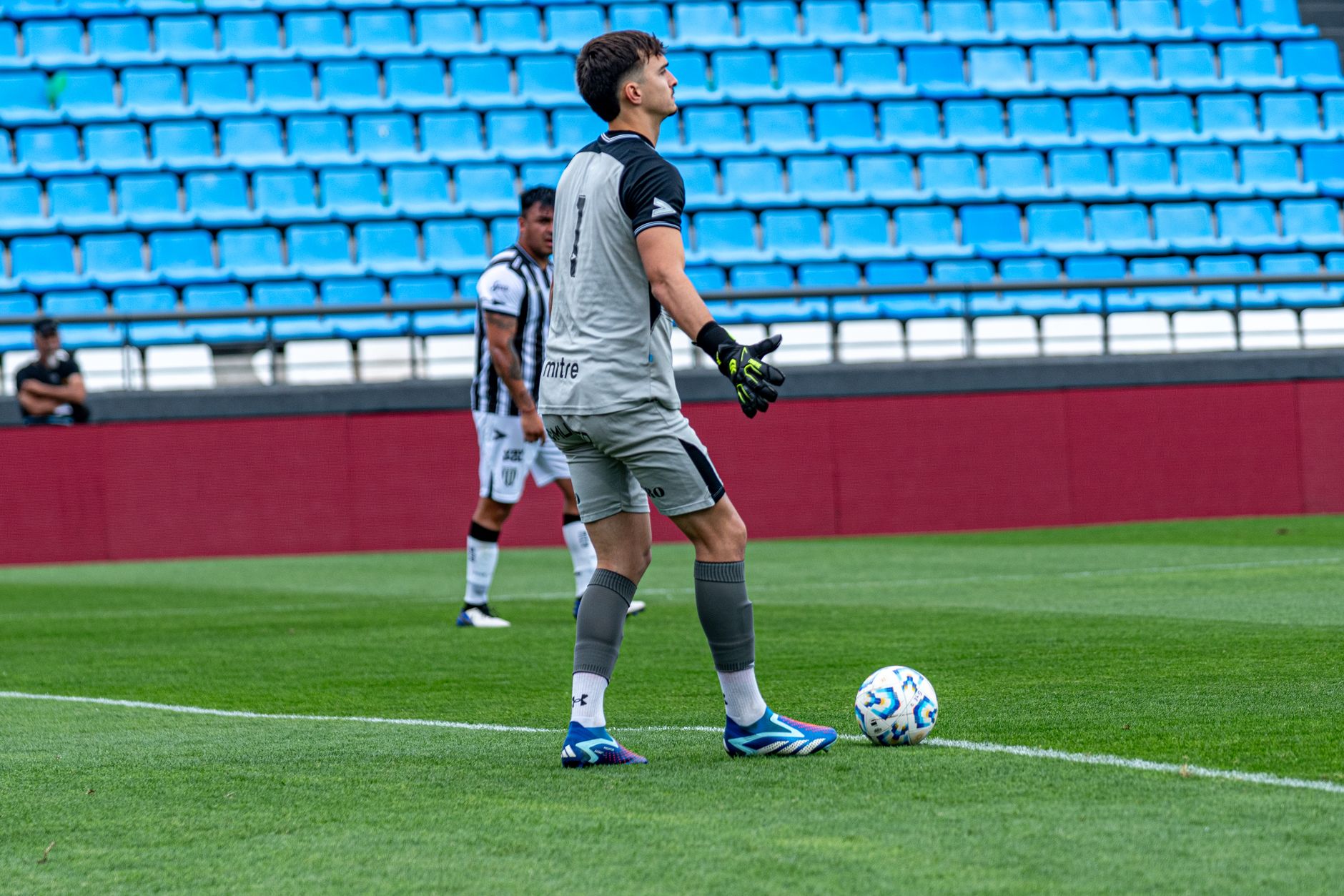 Goalkeeper holding a soccer ball during a match on a bright green field with empty blue seats in the background.