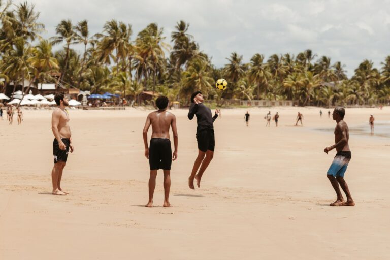 A group of men play soccer on a sandy beach surrounded by palm trees on a sunny day.