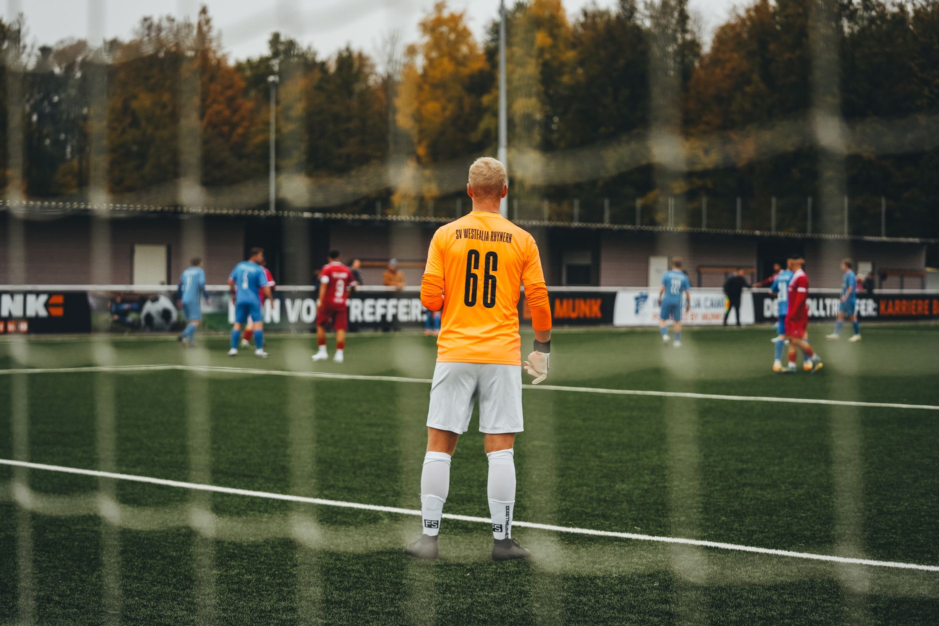 Goalkeeper in orange jersey stands ready during a soccer match on a fall day.