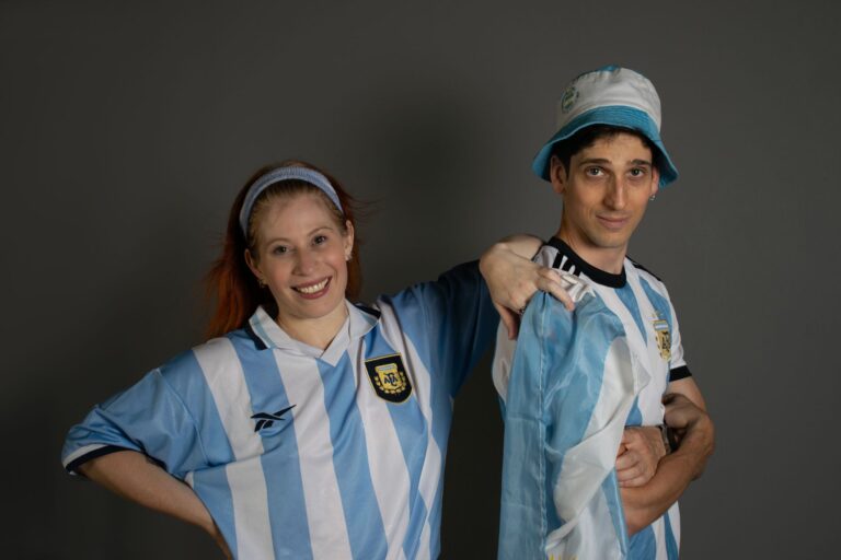 Smiling Argentinian soccer fans wearing team jerseys and a hat, supporting their national team indoors.
