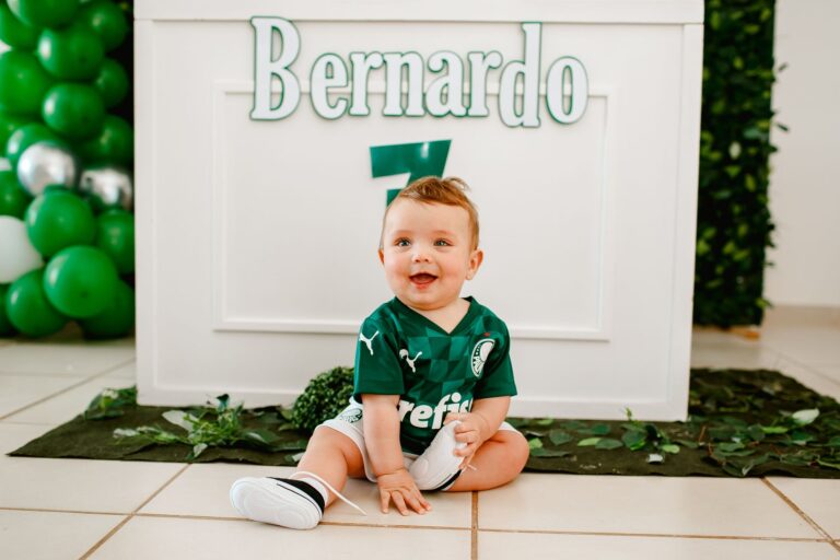 Cute baby in a green jersey sitting indoors surrounded by balloons and greenery.