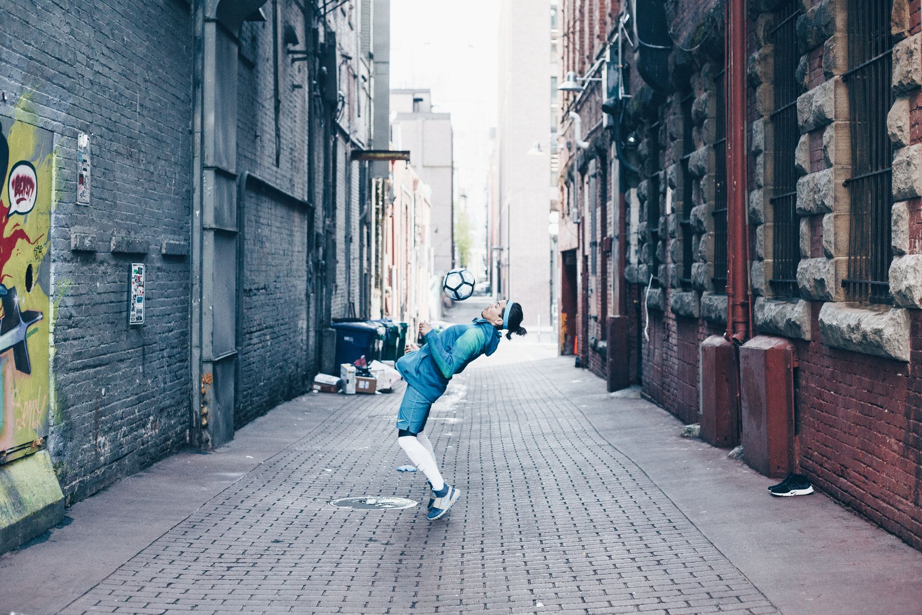 Young man performing soccer tricks in an urban cobblestone alleyway filled with captivating street art.