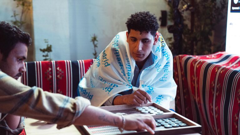 Two men engaged in a game of backgammon, surrounded by colorful textiles, creating a warm indoor atmosphere.