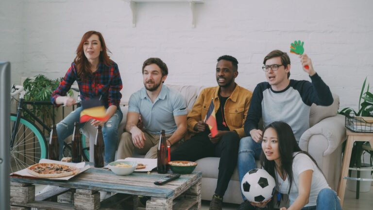 Friends watching a soccer match indoors, waving flags and cheering with snacks and drinks.