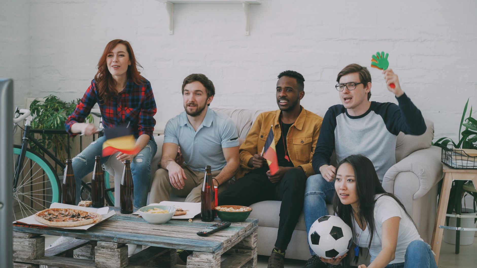 Friends watching a soccer match indoors, waving flags and cheering with snacks and drinks.