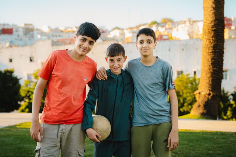 Three boys enjoying a sunny day in Tanger, Morocco, holding a soccer ball in a park setting.