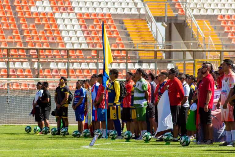 Multiple soccer teams stand in line in a brightly lit outdoor stadium for a ceremony or event.